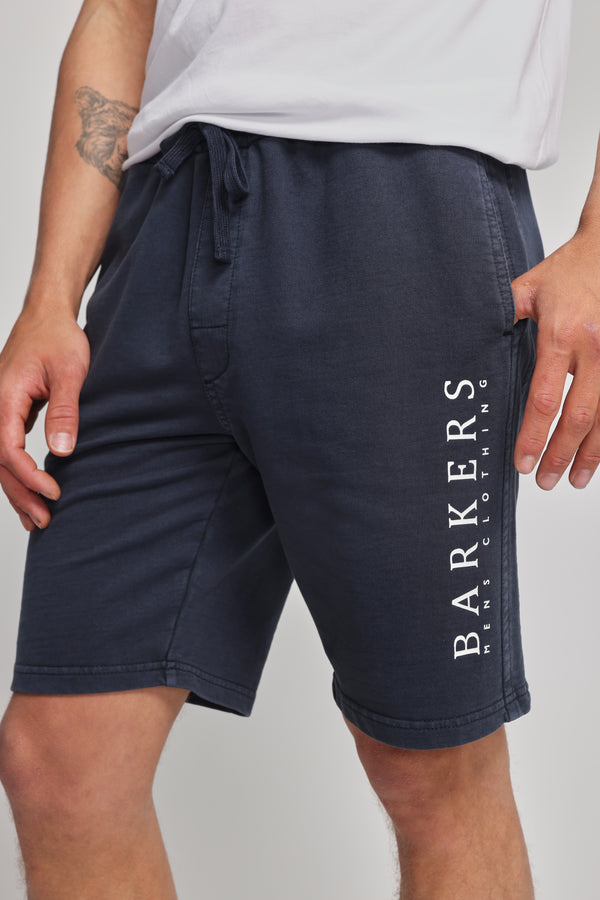 A person wears Barkers Organic Classic Print Track Short in navy, featuring a vertical heritage logo on the left leg, paired with a white t-shirt. Only the lower torso and upper legs are shown.