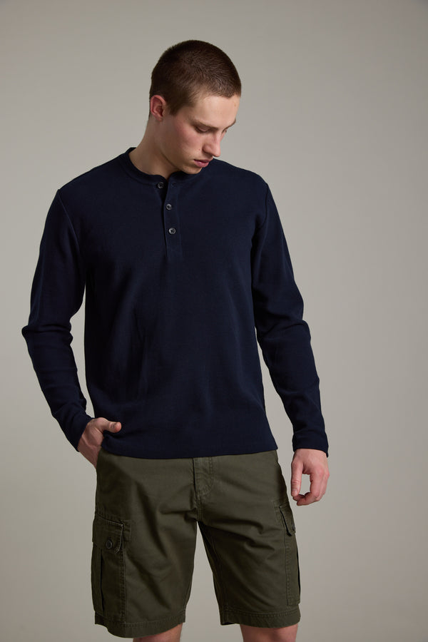 A young man with short hair looks down, one hand in his pocket, wearing the navy Avenue Waffle Henley by Barkers and olive green cargo shorts, standing against a plain gray background.
