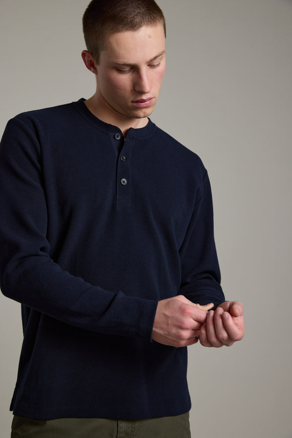 A young man wears the Barkers Avenue Waffle Henley in dark navy—an organic cotton, mid-weight top—and olive green pants, adjusting his cuff against a plain, light gray background.