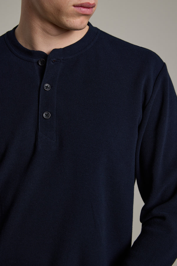 A person is shown from shoulders to mid-torso, wearing the Barkers Avenue Waffle Henley in dark blue organic cotton—a men's mid-weight layer with a three-button collar, facing slightly to the side.
