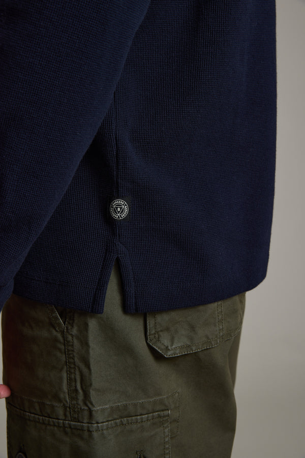 A close-up of a person wearing olive green pocketed pants and the Avenue Waffle Henley by Barkers, a men's mid-weight navy blue organic cotton sweater featuring a small round black and white tag near the hem.