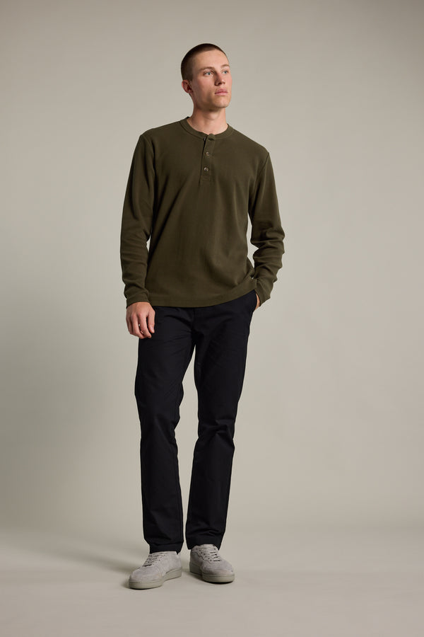 A man stands against a plain light background, wearing the Barkers Avenue Waffle Henley in olive green, paired with dark pants and white sneakers. He has short hair, one hand in his pocket, looking slightly to the side.