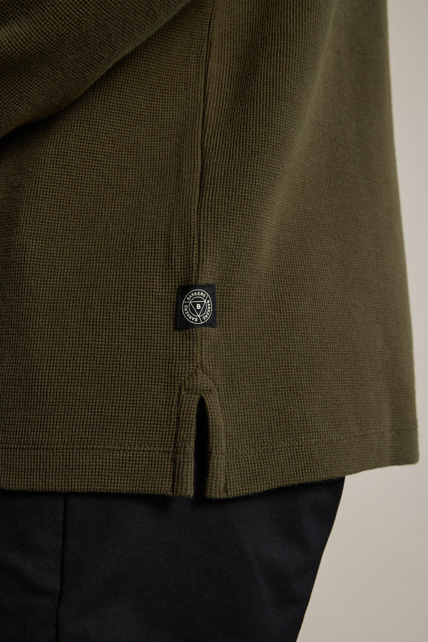 Close-up of the Barkers Avenue Waffle Henley in olive green organic cotton with textured waffle knit, featuring a black and white rectangular label with a circular logo near the hem. Shown paired with black pants.