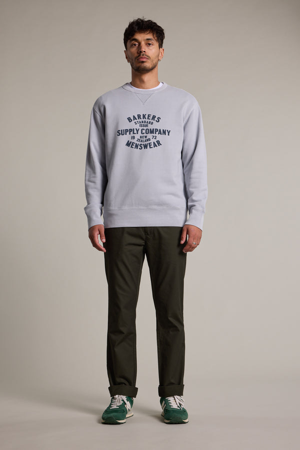 A man faces forward in a Barkers Standard Issue Print Crew sweater in light gray, made from responsibly sourced cotton, styled with dark green pants and green sneakers. He stands against a plain neutral background.