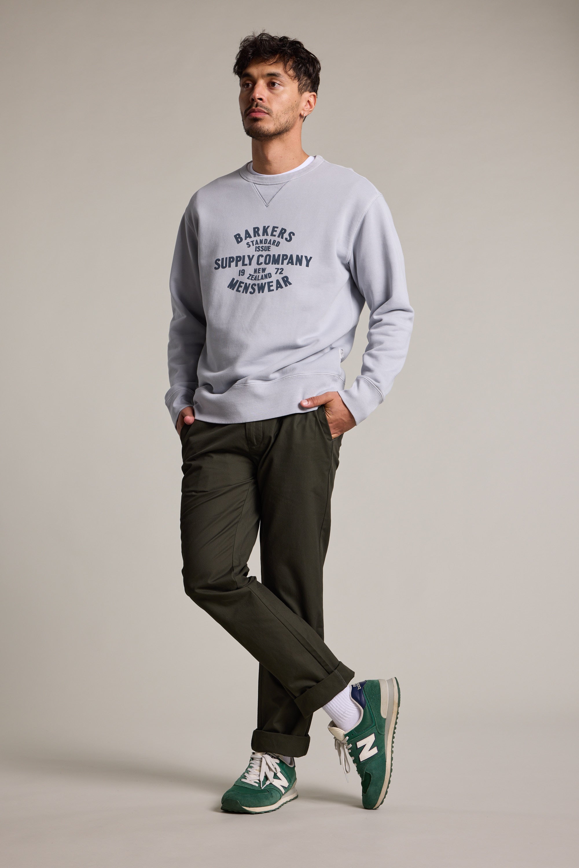A man with short dark hair stands against a plain background wearing a Barkers Standard Issue Print Crew sweater in light gray fleece, dark green pants, and green New Balance sneakers. One hand is in his pocket as he looks slightly to the side.