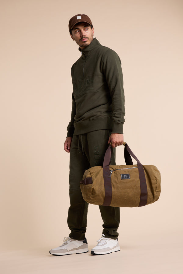 Man in Barkers Heritage 1/4 Zip, white sneakers, brown cap, holding a brown duffel bag.