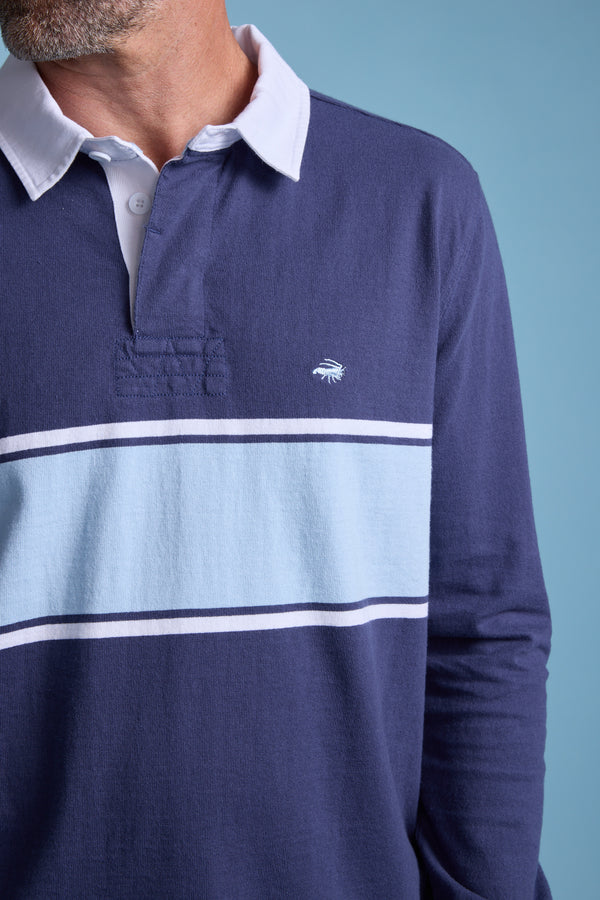 A man wears the Barkers Breaker Rugby Jersey, navy with a white collar, light blue chest stripe bordered by thin white lines, and a small embroidered logo. Made from organic cotton. Light blue background.