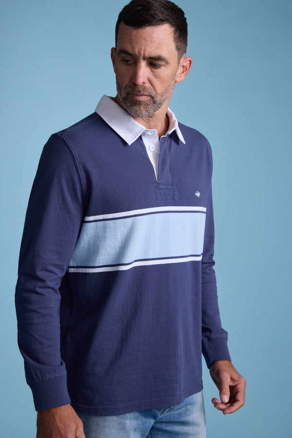 A man with short hair and a beard wears the Barkers Breaker Rugby Jersey, featuring a white collar and horizontal light blue stripe, made from organic cotton, set against a plain light blue background.