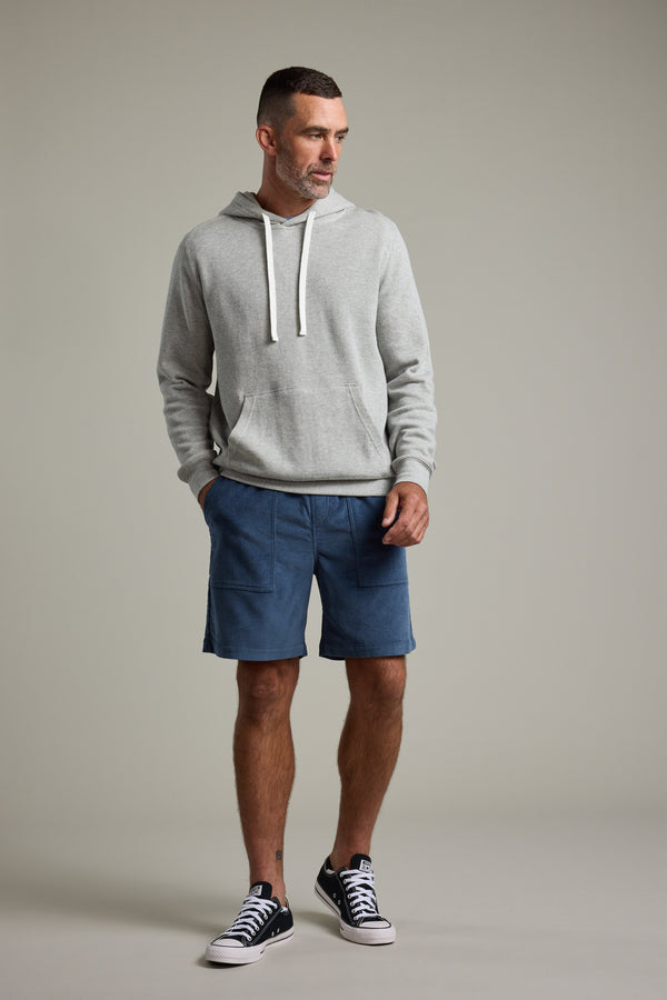 A man with short hair and a beard stands against a plain background wearing the Barkers Inlet Hoody in light gray, blue shorts, and black low-top sneakers with white soles, his hands tucked in the hoody's pockets.