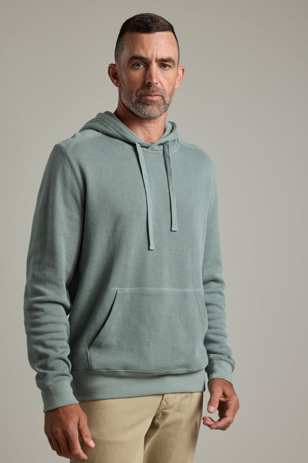 A man with short dark hair and a trimmed beard wears the Barkers Inlet Hoody in light green with beige pants, standing against a plain light gray background.