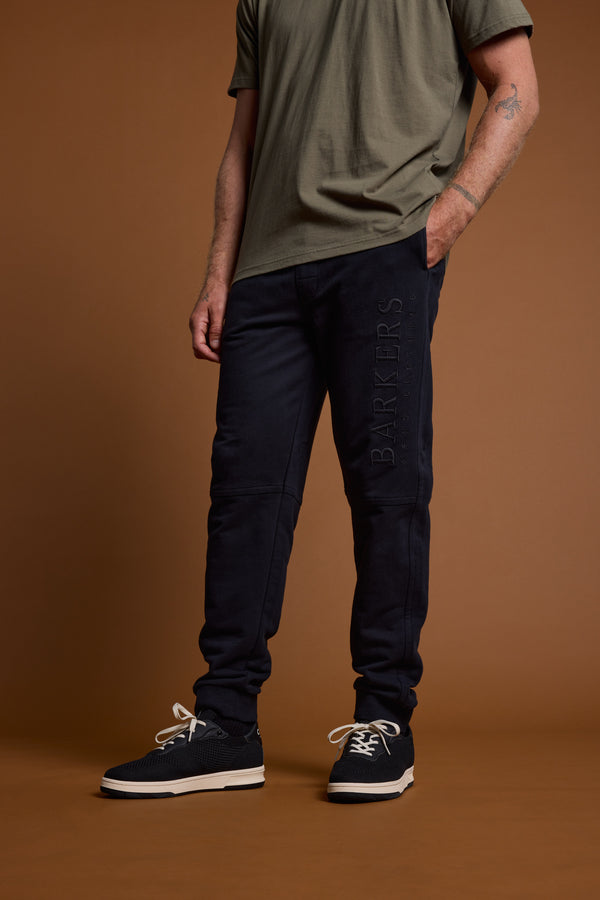 Against a brown background, a person (face not visible) wears a green t-shirt, Barkers Heritage Track Pant by Barkers in dark navy with branded leg text, and black sneakers featuring white soles and laces.