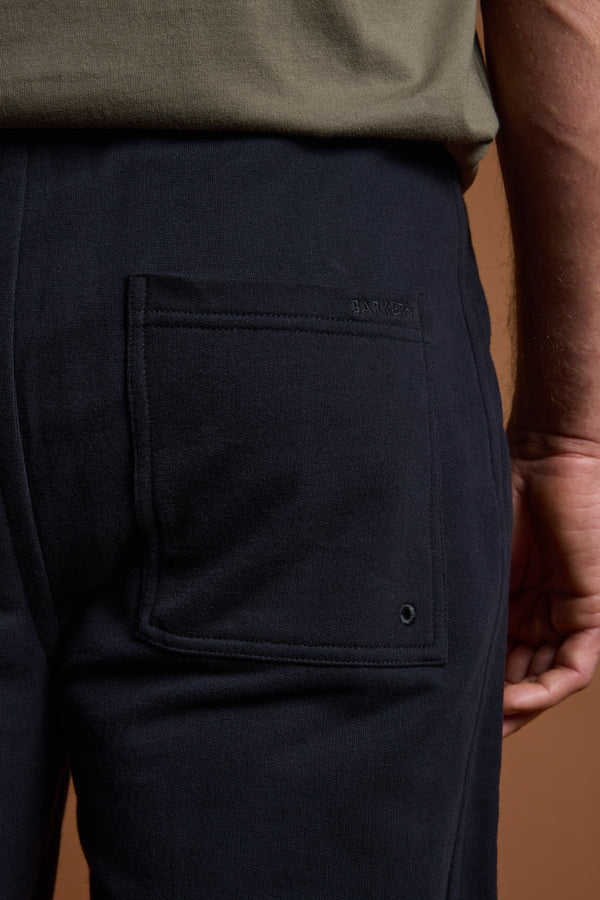 A person wears Barkers Heritage Track Pant in dark navy with a visible back pocket, paired with an olive green shirt. Their right arm rests by their side against a brown background. Brand: Barkers.