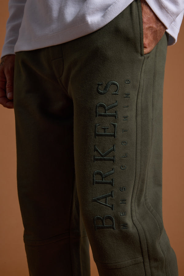 A close-up shows a person wearing olive green Barkers Heritage Track Pant by Barkers, with "BARKERS MENS CLOTHING" embroidered vertically on the left leg, paired with a white long-sleeve shirt and one hand in their pocket.