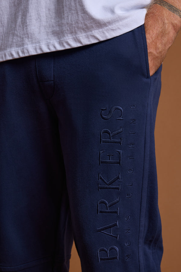 A person wears Barkers Heritage Track Pant in navy, featuring BARKERS MENS CLOTHING embossed vertically on the leg, paired with a partially tucked-in white shirt and one hand in the pocket.