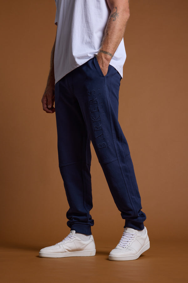 Only the lower half of a person is visible as they stand against a brown background, wearing navy blue Barkers Heritage Track Pants featuring BARKERS embroidered on the leg, paired with a white T-shirt and white sneakers.