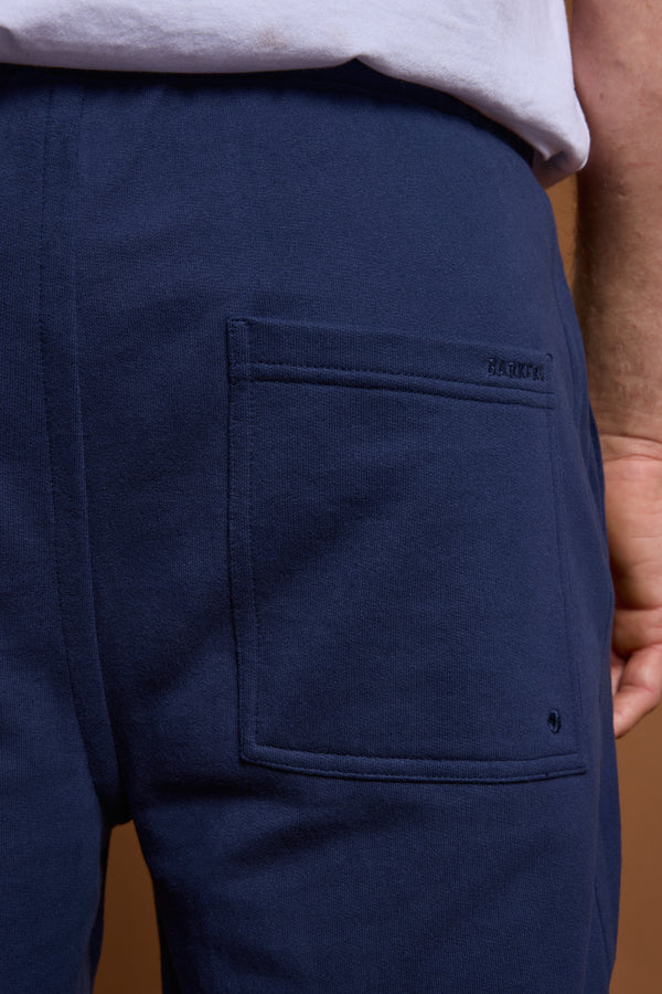Close-up of a person wearing Barkers Heritage Track Pant in navy blue French terry with a square back pocket, partially showing their hand and white shirt. The brand name Barkers is stitched above the pocket.