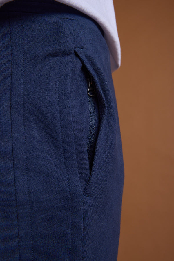 Close-up of a person wearing Barkers Heritage Track Pant in navy blue organic cotton with a zippered pocket, styled with a slightly tucked-in white shirt, by Barkers, against a brown background.