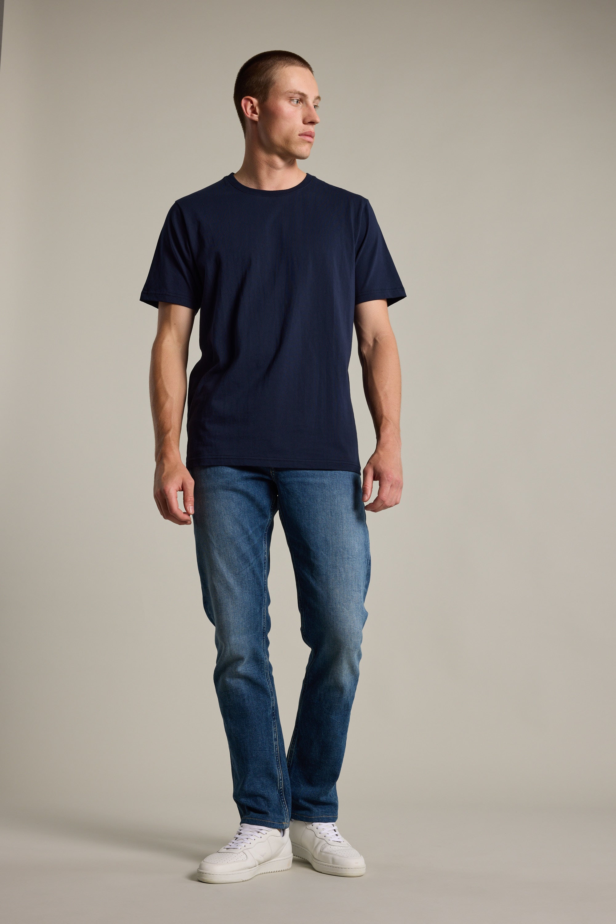 A man wears the Barkers Organic Crew Tee in navy, paired with blue jeans and white sneakers, standing against a plain background. Made from soft organic cotton, this mid-weight tee offers everyday comfort.