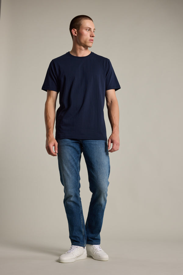 A man wears the Barkers Organic Crew Tee in navy, paired with blue jeans and white sneakers, standing against a plain background. Made from soft organic cotton, this mid-weight tee offers everyday comfort.
