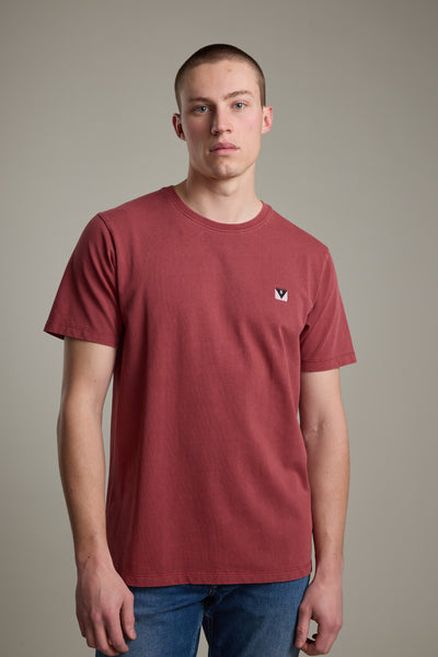 A young man with short hair wears the Barkers Newton Washed Tee in faded red, featuring a small chest logo, paired with blue jeans and standing against a plain light grey background.