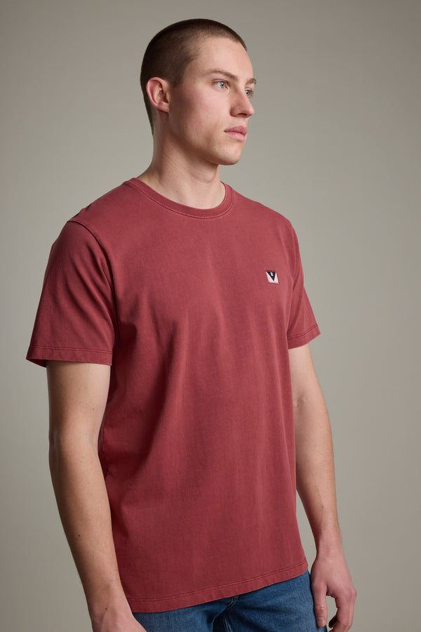 A man with short brown hair models the Barkers Newton Washed Tee in faded red, featuring a small white and black chest logo, as he stands against a plain gray background looking slightly to the side.