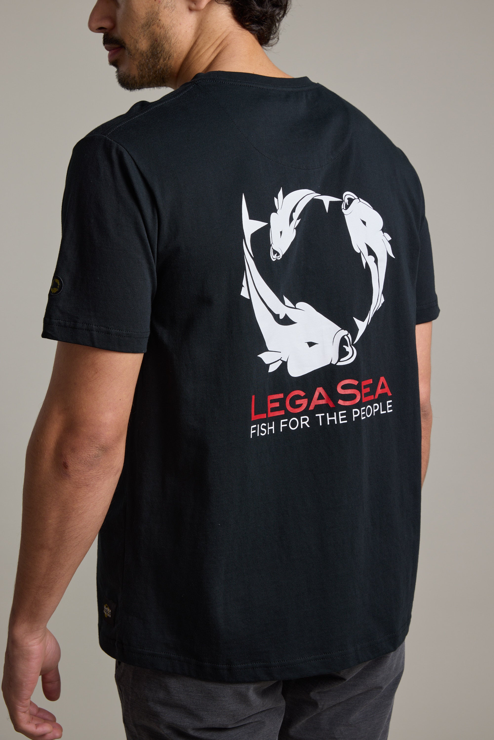 A man wears the Legasea Brand Tee by Legasea, crafted from organic cotton with white fish graphics in a circle and "LEGA SEA FISH FOR THE PEOPLE" on the back, supporting marine conservation in New Zealand.