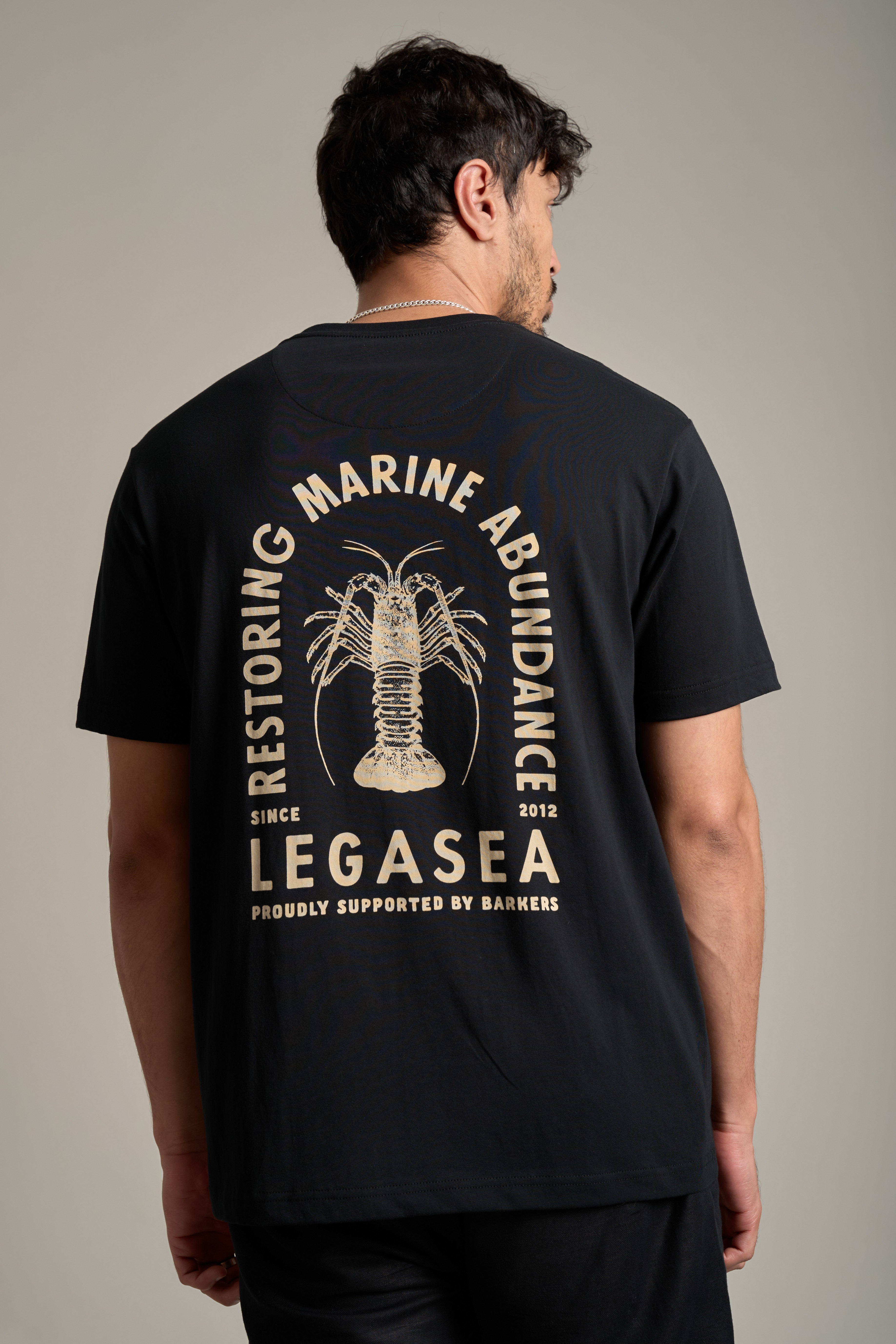 A man wears the Legasea Crayfish Print Tee, an organic cotton shirt with a bold crayfish design and "Restoring Marine Abundance," supporting New Zealand marine conservation since 2012, proudly backed by Legasea.