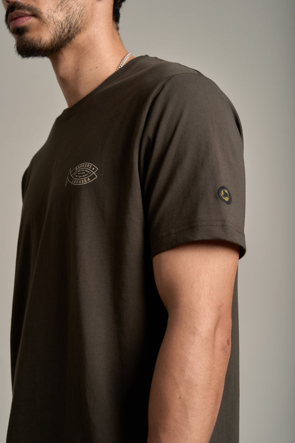 A man wearing the Legasea Crayfish Print Tee, a dark brown organic cotton shirt with a small chest logo and round sleeve patch, stands against a neutral background—showing support for New Zealand marine conservation.