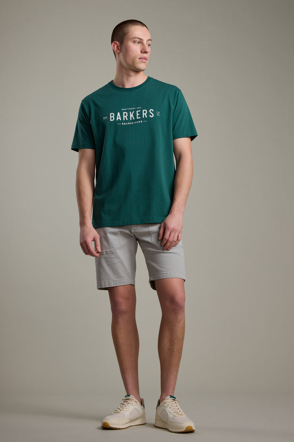 A man stands against a plain background, wearing the Barkers Boardriders Logo Tee in green, paired with light gray shorts and white sneakers with beige soles. He gazes slightly left, maintaining a neutral expression.