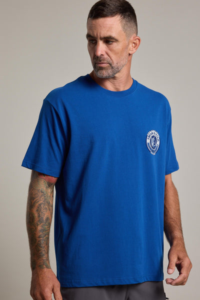 A man with short hair and a beard wears a Swordfish Mojo Logo Tee by Swordfish Mojo in blue organic cotton with a white logo on the left chest. He has a tattoo sleeve on his right arm and looks left against a plain light background.