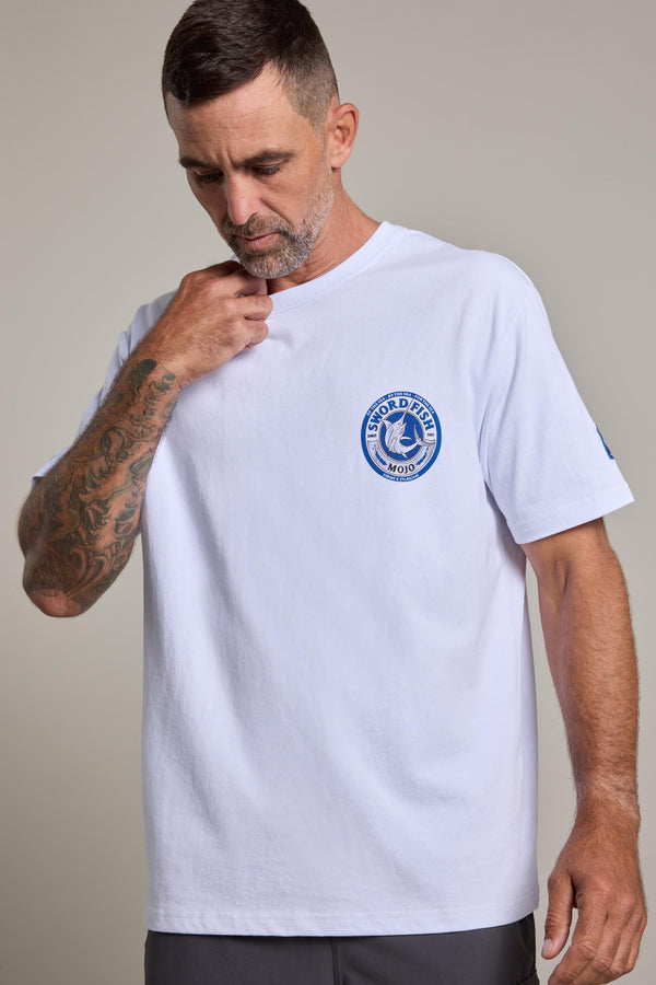 A man with short hair and a tattooed right arm wears a Swordfish Mojo Logo Tee by Swordfish Mojo, featuring a blue circular logo on the chest. He looks down, touching his collar against a plain, neutral background.