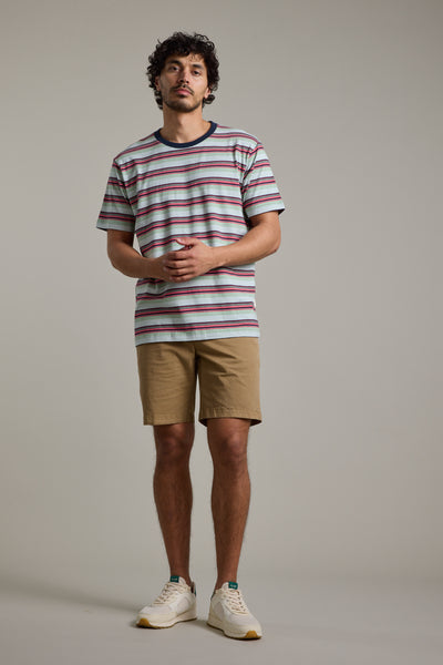 A man with curly hair and a slight beard stands against a plain backdrop, wearing the Barkers Alvin Stripe Tee with a crew neck, tan shorts, and white sneakers, his hands loosely clasped in front of him.