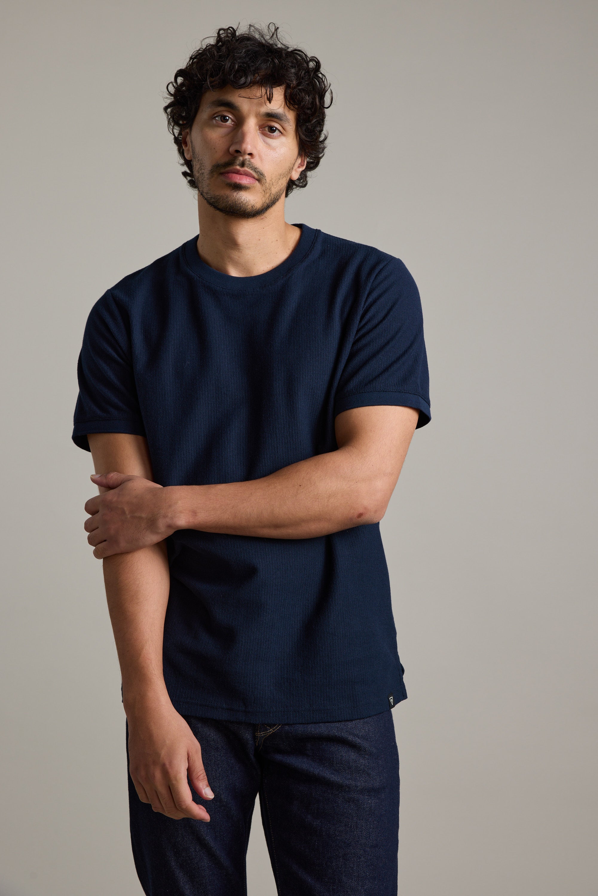 A man with curly dark hair and slight facial hair stands against a plain background, wearing the Barkers Signature Knit Tee in dark blue and dark jeans, with one arm crossed over to hold his other arm above the elbow.