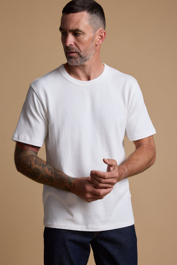 Man with short hair and tattoos wears the Barkers Langwood Tee against a beige background.