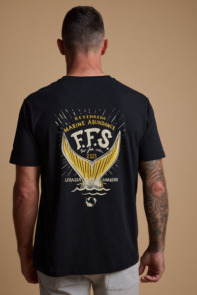 A man with a tattooed right arm wears a black Legasea Tail Print Tee by Legasea, featuring a yellow fish tail graphic and "Restoring Marine Abundance F.F.S for the sake of 2025 LegaSea Barkers" printed on the back.