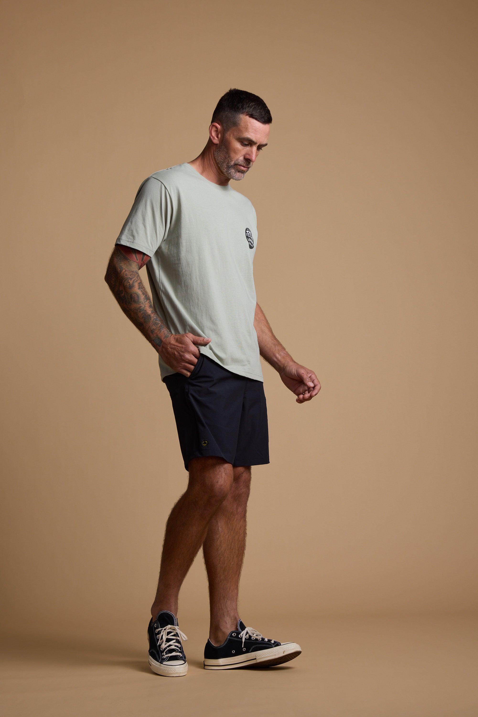 A man with short hair and tattoos wears a light gray T-shirt, Legasea Weekend Short in black, and black sneakers. He stands against a plain beige background, looking down with one hand by his side and the other slightly raised.