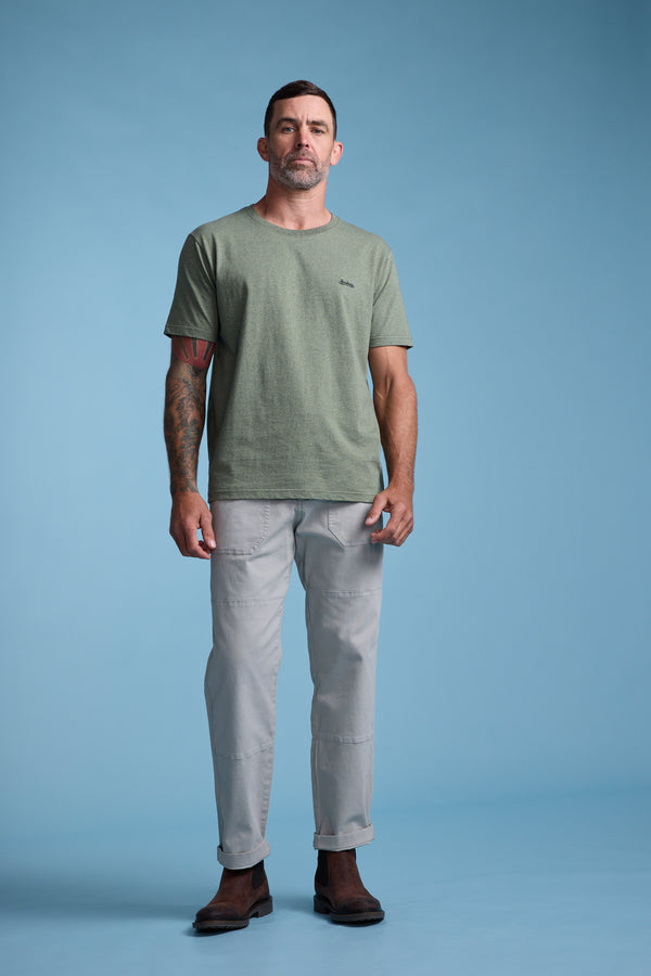 A man with short dark hair, a tattoo on his left arm, and a neutral expression wears the Barkers Bayside Emb Tee in green organic cotton, light gray pants, and brown boots against a plain blue background.
