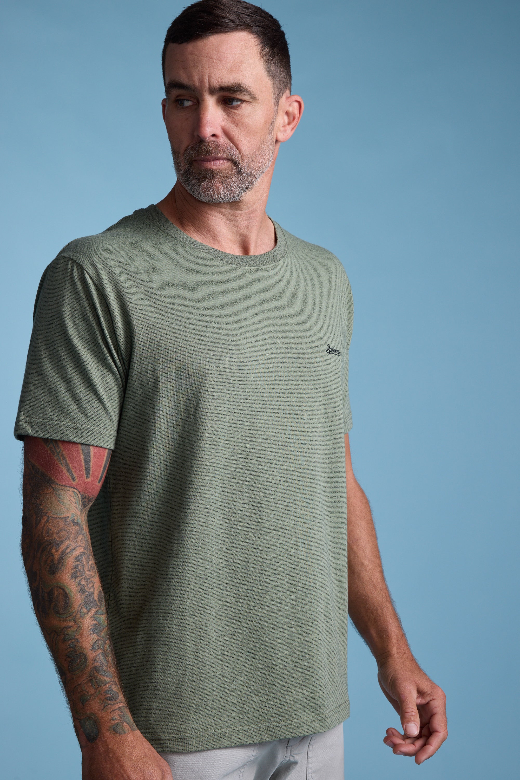 A man with short dark hair, a beard, and a tattoo sleeve wears the Barkers Bayside Emb Tee in olive green with light pants, standing against a light blue background and looking to his left.