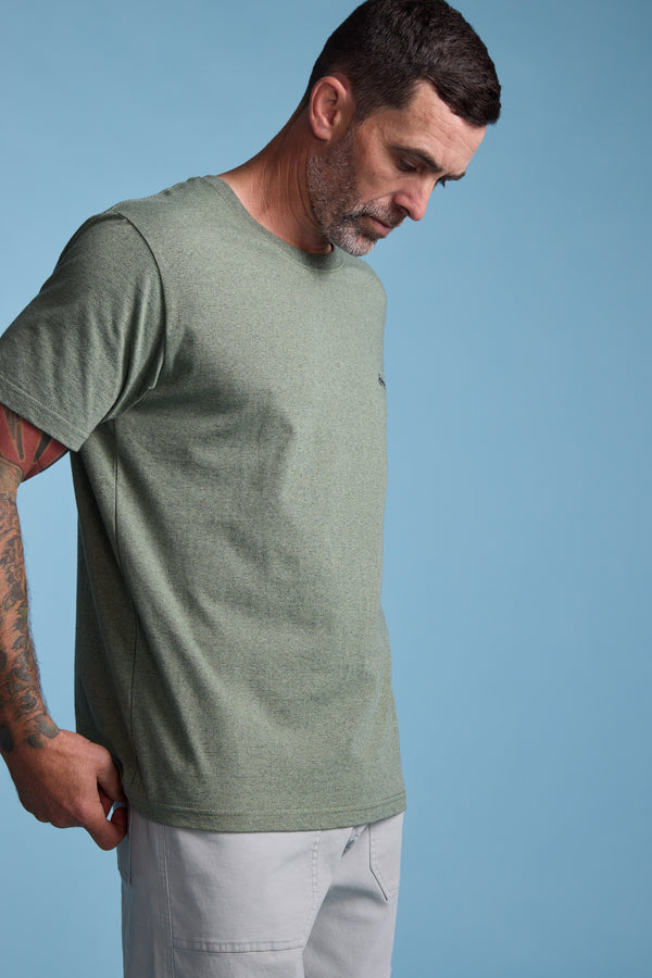 A man with short dark hair and a trimmed beard models the Barkers Bayside Emb Tee in light green, paired with light gray pants. He stands against a light blue background, looking downward, with tattoos visible on his left arm.
