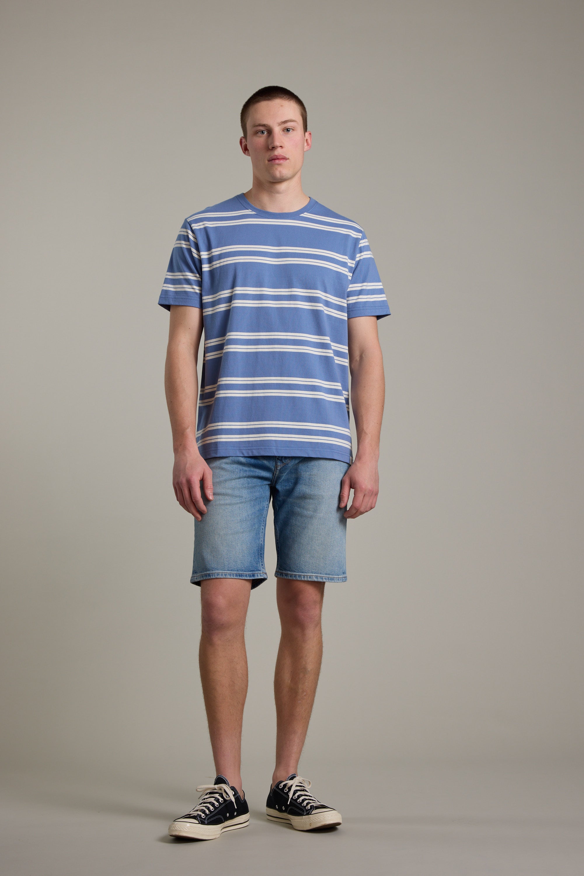 A young man stands against a plain background, wearing the Barkers Northway Stripe Tee—blue and white organic cotton—paired with denim shorts and black-and-white sneakers. He faces forward with a neutral expression, making this tee a summer essential.