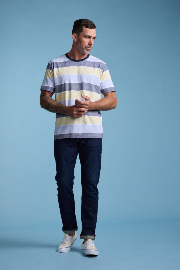 A man with short dark hair and a beard, wearing the Barkers Asbrooke Stripe Tee in pastel shades, dark jeans, and white sneakers, stands against a light blue background looking to his left with his hands clasped.