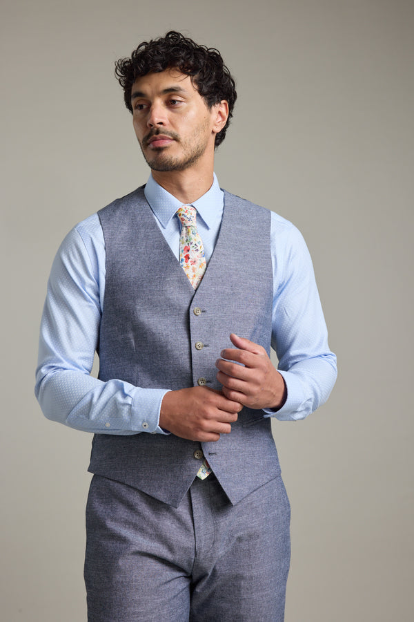 A man wears the Barkers Brighton Cotton Waistcoat over a light blue shirt with matching gray trousers and a floral tie, standing against a plain background while adjusting his vest and looking slightly to the side.
