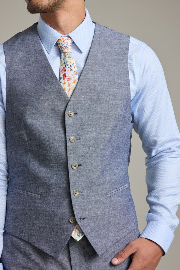 A man wears a Barkers Brighton Cotton Waistcoat over a light blue dress shirt and a colorful floral tie. Only the lower half of his face and upper body are visible, with his right hand resting on his hip.