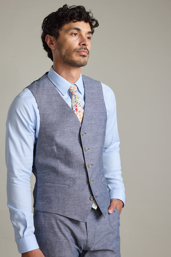 A man stands with one hand in his pocket, wearing Barkers' Brighton Cotton Waistcoat with an adjustable back buckle, a light blue dress shirt, matching gray pants, and a floral tie, gazing slightly to the side against a plain background.