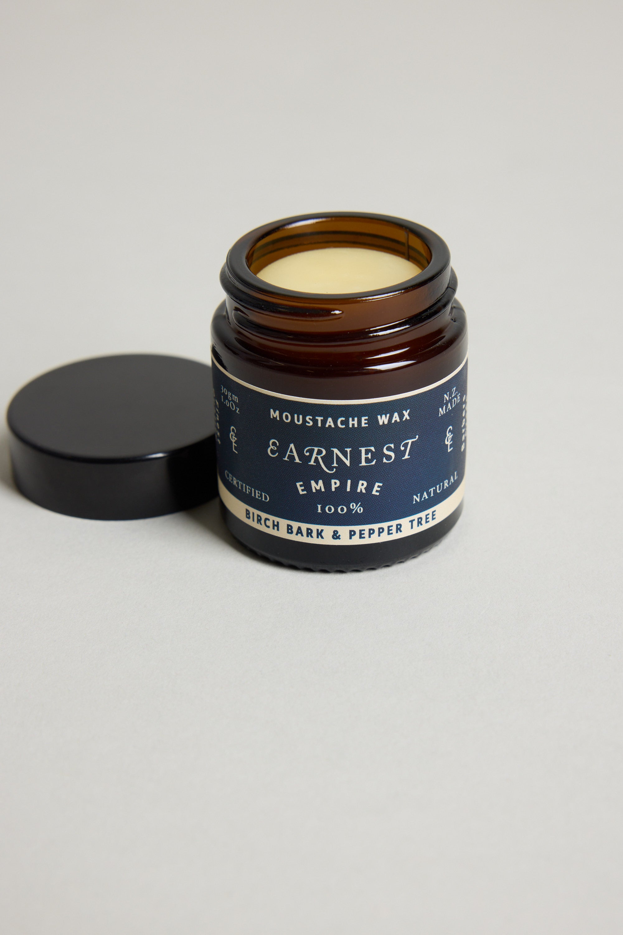 An open jar of Other Earnest Empire Moustache Wax with a dark lid beside it displays "Birch Bark & Pepper Tree" on the label. The light yellow wax inside is 100% natural and provides medium-to-firm hold for your style.