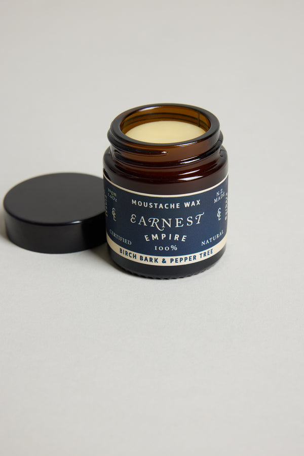 An open jar of Other Earnest Empire Moustache Wax with a dark lid beside it displays "Birch Bark & Pepper Tree" on the label. The light yellow wax inside is 100% natural and provides medium-to-firm hold for your style.