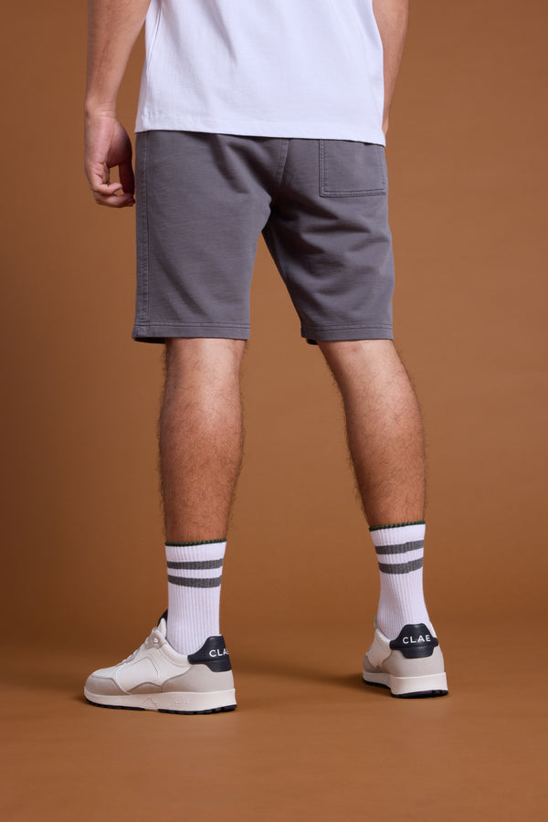 A person stands with their back to the camera, wearing Barkers Organic Classic Print Track Shorts and a white t-shirt, paired with white socks featuring black stripes and white CLAE sneakers, set against a brown background.