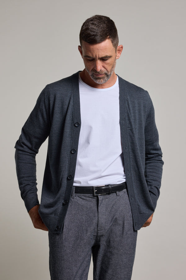 A man wears a white t-shirt, gray trousers, and the Barkers Skyland ZQrx Merino Cardi—crafted from ethically sourced merino wool—standing with hands in pockets, looking down against a plain light background.
