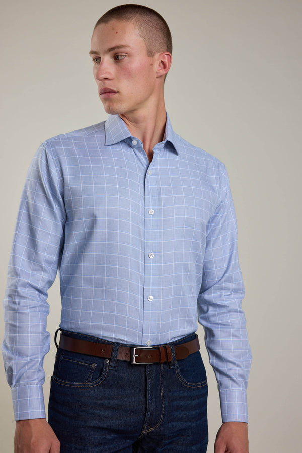 A man wearing the Barkers Tyson Check Business Shirt, a light blue plaid, crease-resistant cotton shirt tucked into dark blue jeans with a brown belt, stands against a plain background looking to his left.