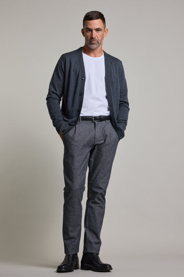 A man with short dark hair and a beard stands against a plain background, wearing the Barkers Skyland ZQrx Merino Cardi over a white t-shirt, gray trousers, and black boots, with his hands in his pockets.